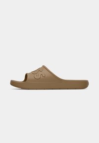 Sandale en caoutchouc brun à bout ouvert, avec un détail texturé, un logo bien visible sur la bride et une semelle intérieure lisse. Design de semelle plate.