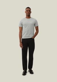 T-shirt cinza de manga curta com decote redondo, combinado com calças pretas de perna reta. O modelo está em pé com as mãos nos bolsos, contra um fundo neutro.