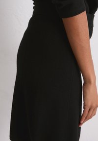 Robe noire en maille vue de côté ; silhouette cintrée avec une texture douce, se terminant au-dessus du genou, présentant une finition lisse et sans coutures.