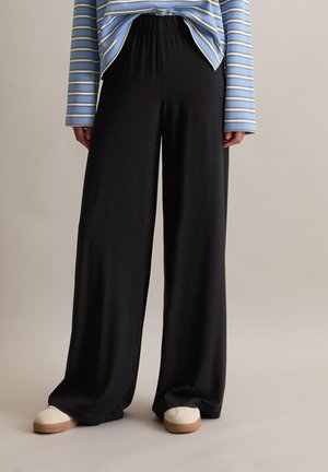 Osoba ubrana w czarne spodnie z szerokimi nogawkami, białe trampki i niebiesko-białą koszulę w paski z długim rękawem, stojąca na tle jednolitego tła.