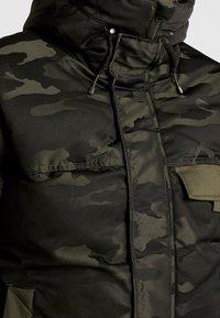 Mężczyzna w ciemnozielonej i czarnej kurtce w kamuflażu z ociepleniem, z wysokim kołnierzem i regulowanymi sznurkami w kapturze.