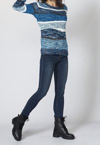 Blå stickad tröja med horisontella ränder i olika nyanser, kombinerad med tajta mörk denimjeans och svarta snörstövlar.