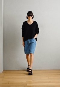 Crni oversized pulover, plava traper suknja i crne sandale. Model stoji na drvenom podu protiv jednostavne sivoj pozadini.