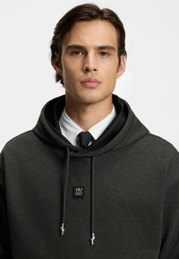 Sudadera con capucha gris oscuro de tela texturizada, con cordones y parche con logo. Se lleva sobre una camisa blanca y corbata negra, con un cuello ancho.