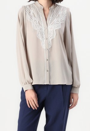 Femme portant un chemisier beige à manches longues avec des détails en dentelle blanche sur le devant et un pantalon bleu marine sur un fond uni.