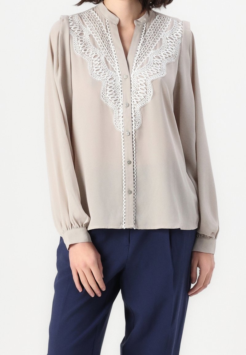 Femme portant un chemisier beige à manches longues avec des détails en dentelle blanche sur le devant et un pantalon bleu marine sur un fond uni.