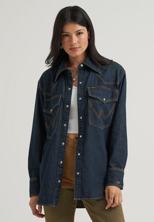 Frau mit dunklen Haaren trägt eine dunkle Jeansjacke über einem weißen Crop-Top und braunen Hosen, stehend vor einem hellgrauen Hintergrund.