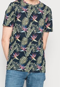 Kortärmad marinblå t-shirt med ett färgglatt tropiskt blommönster som har stora blad och vibrerande fågel av paradiset-blommor. Tillverkad av bomull.