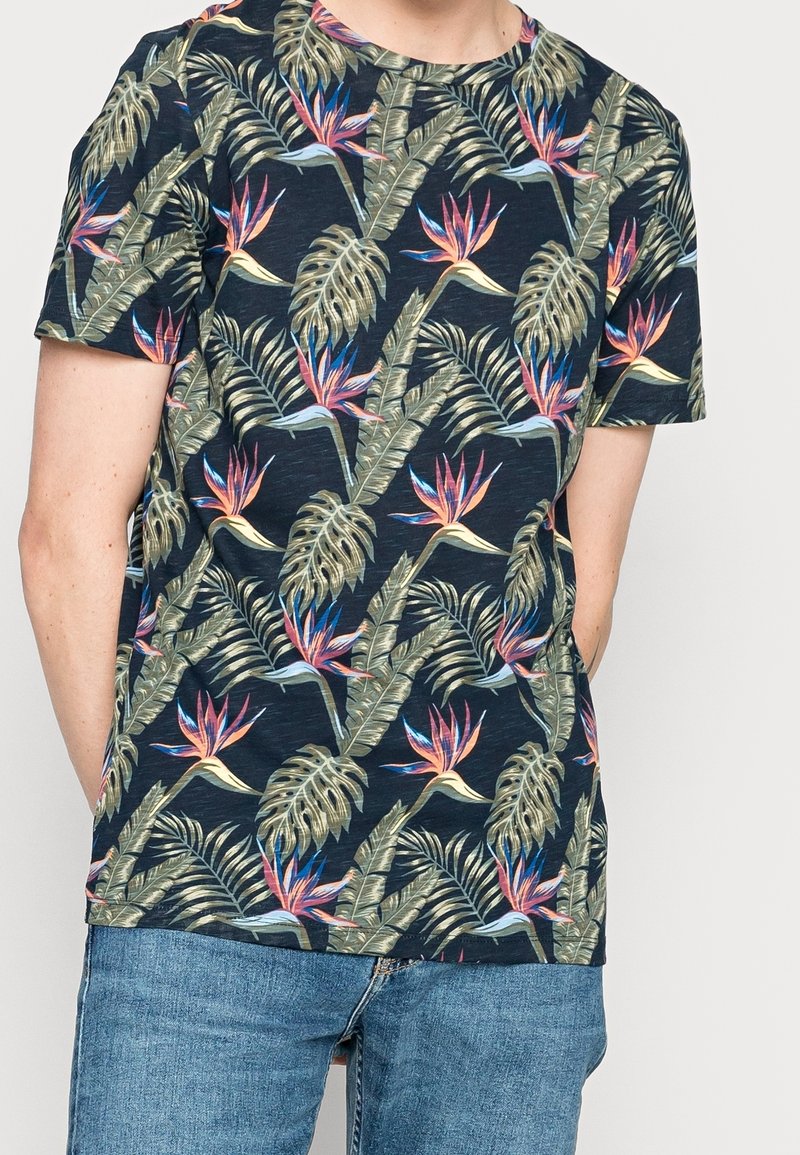 Kortärmad marinblå t-shirt med ett färgglatt tropiskt blommönster som har stora blad och vibrerande fågel av paradiset-blommor. Tillverkad av bomull.