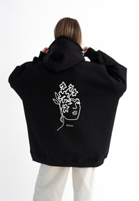 Felpa nera oversize in cotone. Presenta un design di arte lineare bianca di un volto con accenti floreali sulla schiena. Cappuccio attaccato.