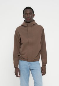 Άνδρας που φοράει καφέ μπουφάν με κουκούλα, ελαστικό τελείωμα και μανσέτες, σε συνδυασμό με ανοιχτό μπλε τζιν, στέκεται μπροστά σε απλό λευκό φόντο.