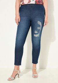 Tmavě modré džíny s těsným střihem, které mají texturované horizontální linie v pase a třpytivý design obličeje na levé nohavici.