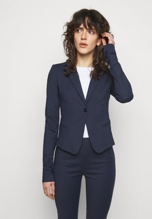 Navy fitted blazer with one button closure, notch lapels, and long sleeves. Paired with matching tailored pants and a white top underneath.