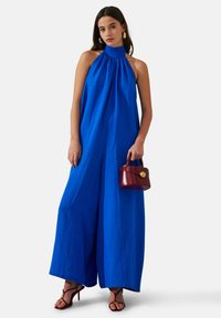 Combinaison bleu à bretelles avec encolure froncée, design à jambes larges, tissu léger. Associée à un petit sac à main rouge et des talons à lanières.