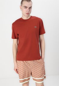 Tricou roșu din bumbac cu guler rotund și un mic logo verde pe piept, asortat cu pantaloni scurți cu model în dungi portocalii, albe și roșii.