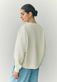 Hellbeige übergroßer Sweatshirt mit Rundhalsausschnitt und überschnittenen Schultern, langen Ärmeln und weichem Material. Sichtbare blaue Streifenhose.