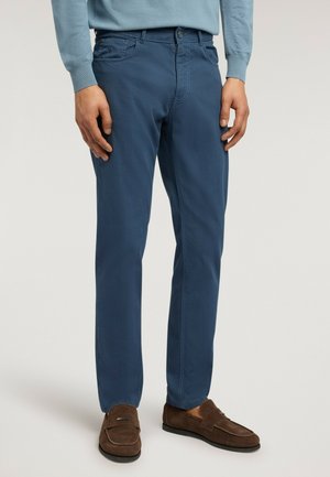 Mand iført blå slim-fit bukser og brune ruskindslædermokkasiner, stående mod en ensartet lys baggrund.