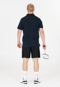 Marinblå pikétröja, svarta träningsshorts, vita strumpor och sneakers; håller en badmintonracket med texturerat handtag, vänd bort.