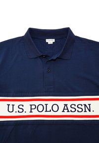 Tummansininen polo-paita, jossa on ribbitetty kaulus, kaksi nappia ja valkoinen vaakasuora raita punaisilla reunoilla, jossa lukee "U.S. POLO ASSN."