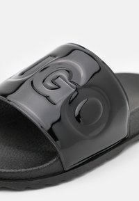 Svart slide-sandal med texturerad fotbädd och glänsande övre rem med stora präglade bokstäver "G" och "O".