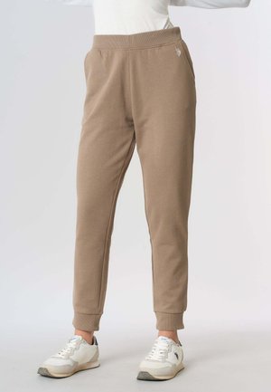 Pantaloni jogger beige con caviglie risvoltate indossati da una persona in piedi, abbinati a sneakers bianche con dettaglio logo nero.