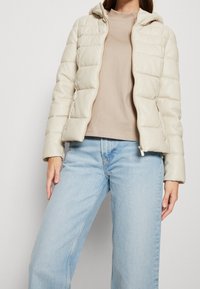 Polstret cremet jakke med hætte, der har lynlås og sidelommer; båret over en beige top og lyseblå jeans.