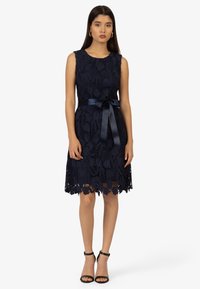 Robe sans manches en dentelle marine avec motif floral, ourlet festonné et ceinture en satin. Assortie à des sandales à talons noires.