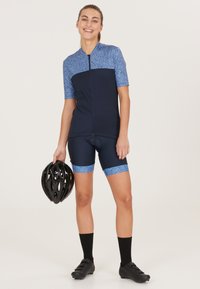 Tenue de cyclisme à manches courtes, corps bleu marine avec des accents bleus à motifs. Porte un casque noir, un short jusqu'aux genoux et des chaussettes noires.