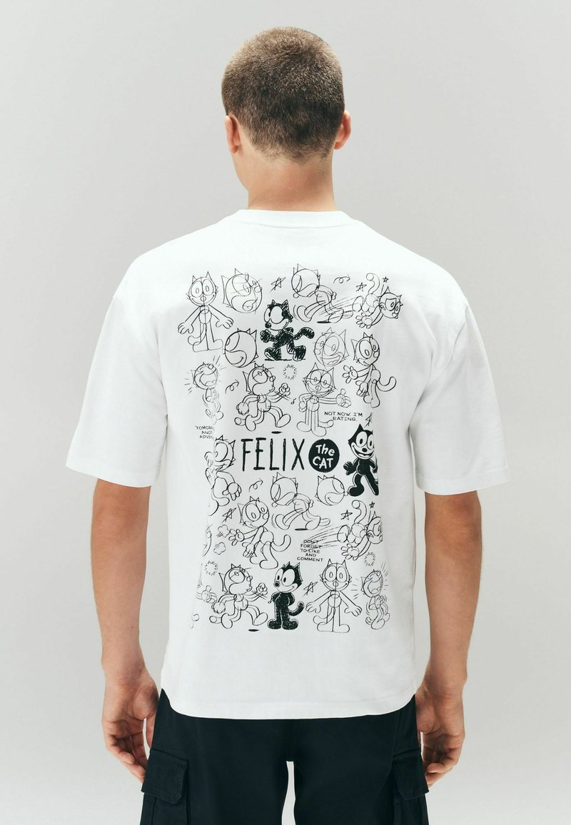 Uomo che indossa una maglietta bianca con schizzi a fumetti in bianco e nero di Felix the Cat e testo sul retro, in piedi davanti a uno sfondo semplice.