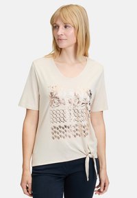 Beige kortærmet t-shirt med dyb udskæring, prydet med et metalisk blomstermønster og strukturelle mønstre. Bundet knude detalje i kanten.