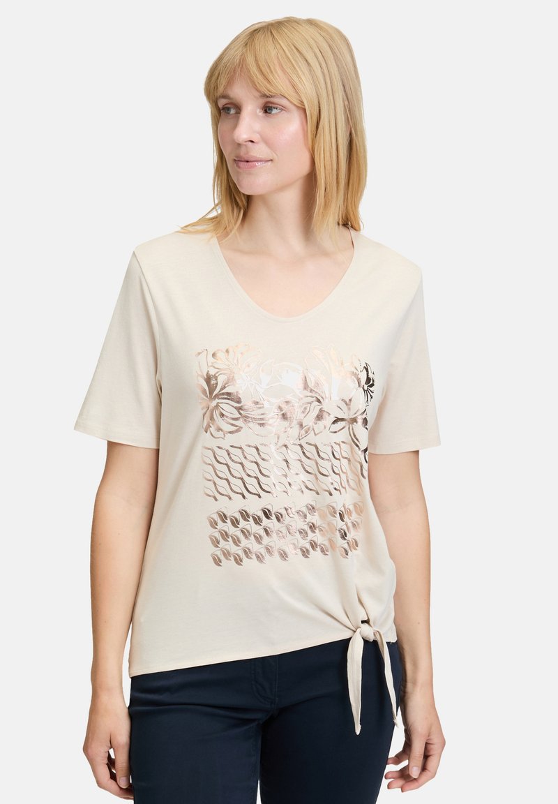 Beige kortærmet t-shirt med dyb udskæring, prydet med et metalisk blomstermønster og strukturelle mønstre. Bundet knude detalje i kanten.
