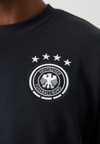Svart hettegenser med et rundt brodert merke som har teksten "DEUTSCHER FUSSBALL-BUND" og et design av en dobbelhodefugl, supplert med fire hvite stjerner over.