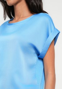 Světlomodrá halenka s hladkou, saténovou texturou, kulatým výstřihem a krátkými rukávy s ohrnutými lemy. Jednoduchý, elegantní design bez vzorů.