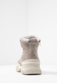 Bottine en suède gris avec un col pelucheux, semelle texturée blanche et fermeture éclair sur le côté. Caractéristiques d'un talon renforcé et d'une boucle décorative à l'arrière.
