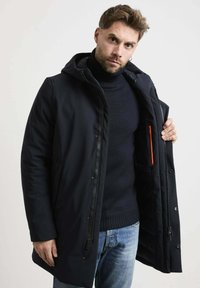 Marineblauwe gewatteerde parka met een rits- en drukknoopsluiting, voorzien van een fleece-gevoerde kraag en een binnenzak met oranje details.