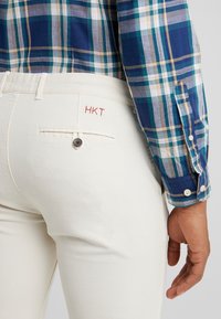 Pantalon en coton beige avec une poche arrière, fermeture par bouton et broderie rouge "HKT", associé à une chemise à manches longues à carreaux bleus.