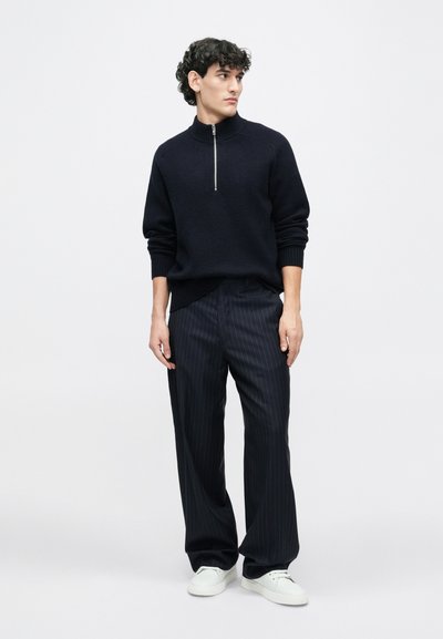 Black knit half-zip sweater, ribbed texture, paired with dark pinstriped trousers and white sneakers. Model stands against a plain background.