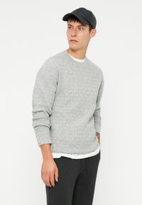 Pull en tricot gris avec un motif texturé, col rond, manches côtelées, porté sur une chemise blanche, associé à un pantalon gris foncé et une casquette noire.