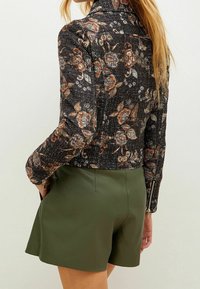 Mønstret sort jakke med blomsterdesign og tekstur, med lange ærmer og lynlåslukning ved håndleddene. Sammen med olivengrønne, højtaljede shorts.