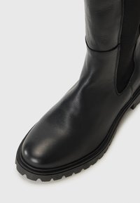 Gros plan sur une botte en cuir noir avec une surface lisse, un panneau latéral élastique et une semelle robuste, sur fond blanc uni.