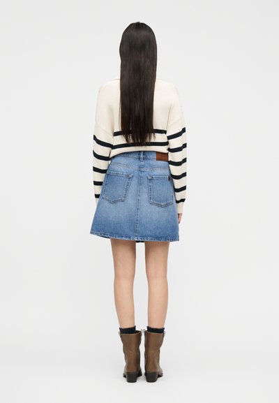 Γυναίκα με μακριά σκούρα μαλλιά, ντυμένη με κρεμ πουλόβερ με μαύρες ρίγες, μπλε τζιν φούστα και καφέ μποτάκια μέχρι τον αστράγαλο, όρθια με την πλάτη γυρισμένη.