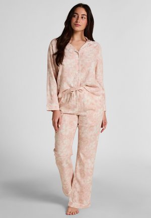 Vrouw staat blootsvoets, draagt een lichtroze pyjamahandschoen met lange mouwen, knoopjes en bloemmotief, en een bijpassende broek met aantrekkoord, tegen een effen achtergrond.