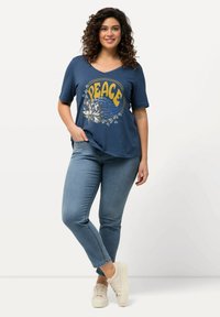 T-shirt in cotone blu con scollatura a V e maniche corte. Presenta una grafica con la scritta "PACE" in giallo, accentuata da illustrazioni floreali. Abbinata a jeans chiari e sneakers beige.
