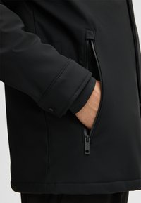 Czarna kurtka softshell z zamykaną na zamek kieszenią i ściągaczami na rękawach. Charakteryzuje się gładką fakturą i aerodynamicznym krojem dla nowoczesnego wyglądu.