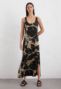 Abito lungo floreale in beige con motivi a foglie nere, caratterizzato da un colletto a girocollo e spacco laterale. Abbinato a sandali neri piatti.