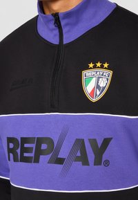 Sweat-shirt noir et violet à fermeture éclair quart, logo Replay FC avec trois étoiles, un blason aux couleurs du drapeau italien et une fleur blanche.