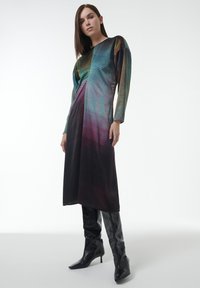 Lang, getailleerde jurk met een gradientpatroon in teal, paars en zwart. Lange mouwen en een ronde halslijn. Gecombineerd met zwarte kniehoge laarzen.