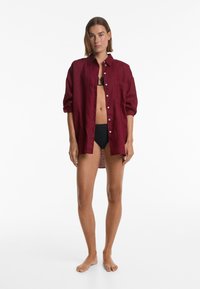 Camicia in lino borgogna con bottoni, maniche arrotolate, taschino frontale e un orlo posteriore leggermente più lungo. Indossata su un bikini nero.