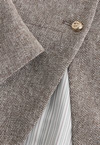 Reverul sacoului cu model chevron maro și alb, cu nasture auriu răsucit și o privire rapidă către o țesătură albă cu dungi verticale subțiri dedesubt.