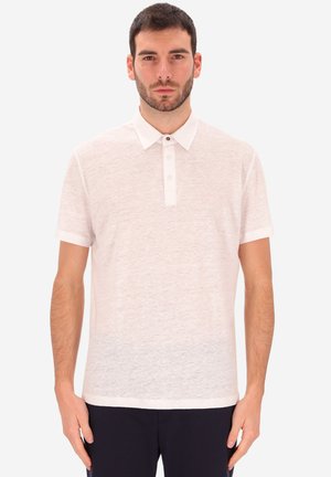 Uomo con capelli corti e scuri e barba, che indossa una polo bianca a maniche corte e pantaloni scuri, in piedi davanti a uno sfondo chiaro e uniforme.